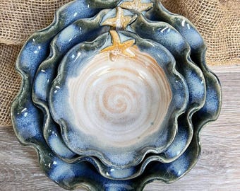 Pode incluir: Um conjunto de três taças de cerâmica encaixáveis com bordas onduladas e festonadas. As taças apresentam um esmalte degradê que transita do azul profundo nas bordas para o bege areia no centro, decoradas com um pequeno detalhe de estrela.