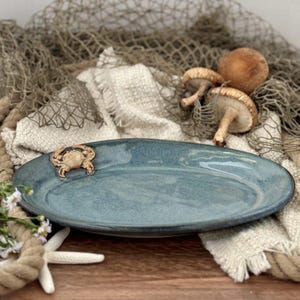 May include: A blue oval ceramic platter with a crab-shaped accent. The platter is approximately 30 cm long and has a rustic, handmade appearance. The background includes mushrooms, a fishing net, and a starfish.