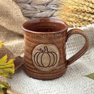May include: A speckled brown ceramic mug with a cylindrical body and a curved handle. The exterior features a circular medallion on the front displaying a carved line drawing of a pumpkin. The surface has a textured, earthy finish throughout.