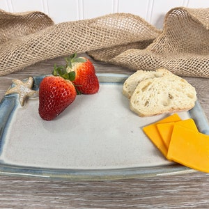 May include: A rectangular ceramic serving tray with a light gray center and a blue-toned rim, featuring a small starfish accent on the top left corner. The tray holds two fresh red strawberries, two slices of crusty bread, and three slices of cheddar cheese.