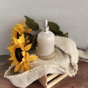 May include: A speckled ceramic soap dispenser with a silver pump sits on a woven beige cloth. Bright yellow sunflowers with dark centers and green leaves are arranged to the left. The dispenser rests on a small, round, light brown base.