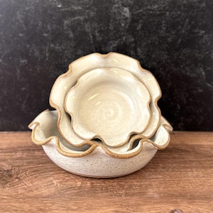 May include: Set of three ceramic bowls with a speckled, off-white glaze and a wavy, ruffled rim. The bowls are nested, with the largest bowl at the bottom and the smallest at the top. The rims have a brown trim. The bowls are on a wooden surface.