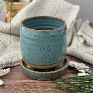 May include: A cylindrical ceramic planter with a speckled teal blue glaze and a natural brown clay base. The planter features horizontal ridges along the exterior and sits on a matching circular saucer with a raised rim and a brown clay edge.