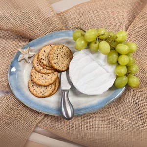 May include: A blue and white ceramic serving plate with a starfish design holds crackers, a knife, a wheel of brie cheese, and a bunch of green grapes. The plate sits on a burlap cloth.