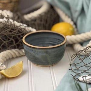 May include: A handmade ceramic bowl with a blue-grey glaze and a tan rim. A lemon wedge rests beside the bowl on a white surface. The background includes a fishing net, rope, and a whole lemon.
