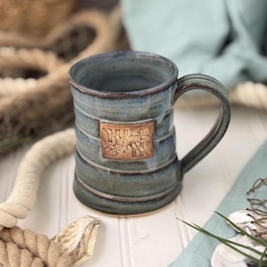 May include: A ceramic mug with a blue and grey glazed finish featuring horizontal ridges along the body. A rectangular clay emblem is centered on the front, displaying a relief design of the Maryland state flag with its distinct heraldic patterns.