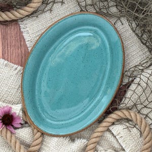 May include: An oval ceramic platter with a glossy turquoise glaze and dark speckles throughout. The platter features a raised rim and a smooth, reflective surface. The edges of the ceramic piece show a natural, unglazed brown clay tone.