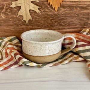 May include: A speckled white ceramic mug with a brown base and a curved handle. The mug sits on a colorful striped cloth. The background includes wooden fall-themed decorations.