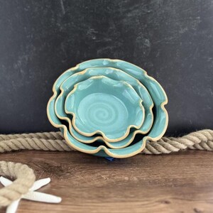 May include: A set of three turquoise ceramic bowls with a ruffled edge. The bowls are nested, with a spiral design in the center. The rims have a light brown trim. The bowls are on a wooden surface with a rope and a starfish.