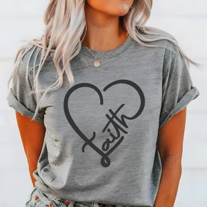 Peut inclure: Un t-shirt gris chiné à manches courtes avec une impression graphique noire représentant une forme de cœur combinée au mot faith écrit en police cursive. Le t-shirt a une encolure ronde et des manches retroussées, offrant un aspect décontracté.