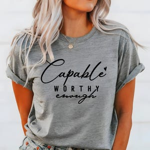 May include: Gray short-sleeved t-shirt with black text that reads "Capable Worthy Enough" with a small heart symbol.