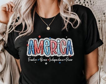 Peut inclure: T-shirt noir avec un graphique "America" en rouge, blanc et bleu avec des pois blancs. Le graphique est entouré de petites étoiles rouges. Le texte "Freedom Brave Independence Power" est sous le graphique.