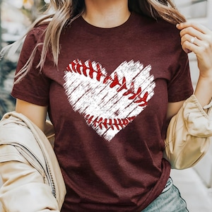 Puede incluir: Una camiseta color burdeos con un gr&aacute;fico blanco en forma de coraz&oacute;n de una pelota de b&eacute;isbol con costuras rojas.