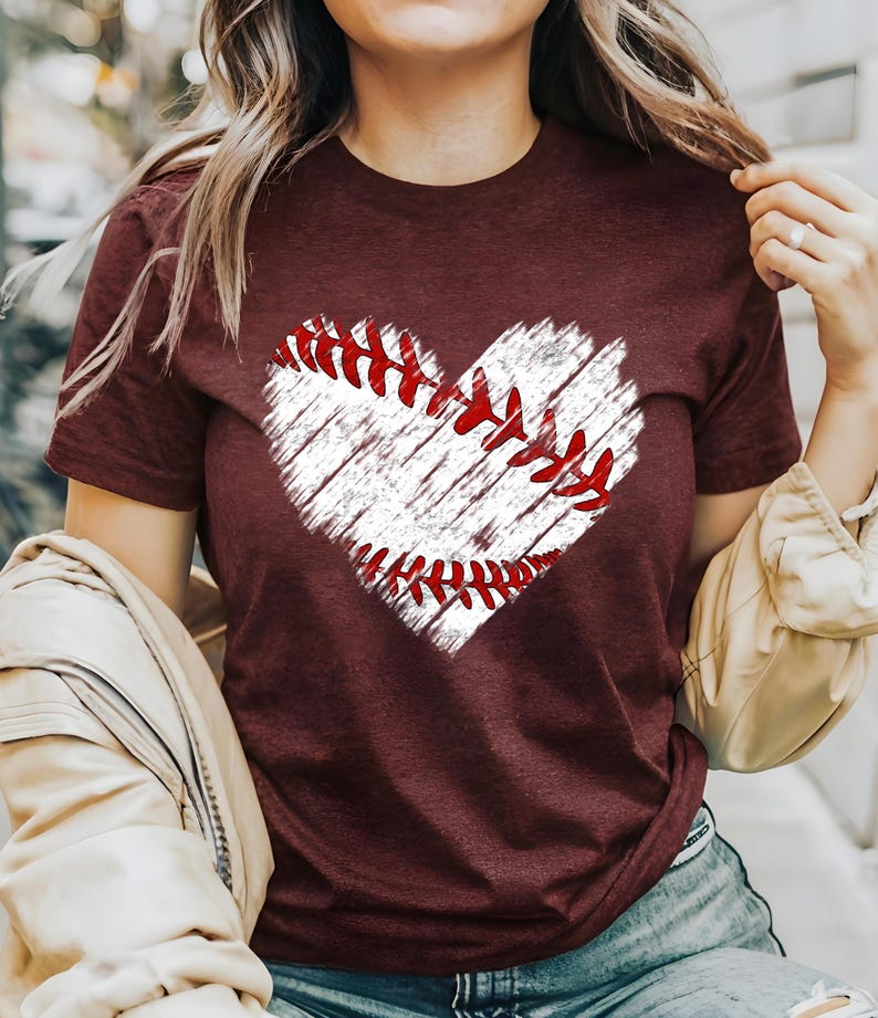 Puede incluir: Una camiseta color burdeos con un gr&aacute;fico blanco en forma de coraz&oacute;n de una pelota de b&eacute;isbol con costuras rojas.