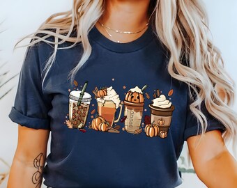 Puede incluir: Una camiseta azul marino con un gráfico de cuatro bebidas con temática otoñal. Las bebidas son un latte de especias de calabaza, un café helado de especias de calabaza, un batido de especias de calabaza y un chocolate caliente de especias de calabaza. El gráfico también incluye hojas de otoño, calabazas y palitos de canela.