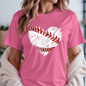 Puede incluir: Una camiseta rosa con un gr&aacute;fico de b&eacute;isbol blanco en forma de coraz&oacute;n. El b&eacute;isbol tiene costuras rojas.