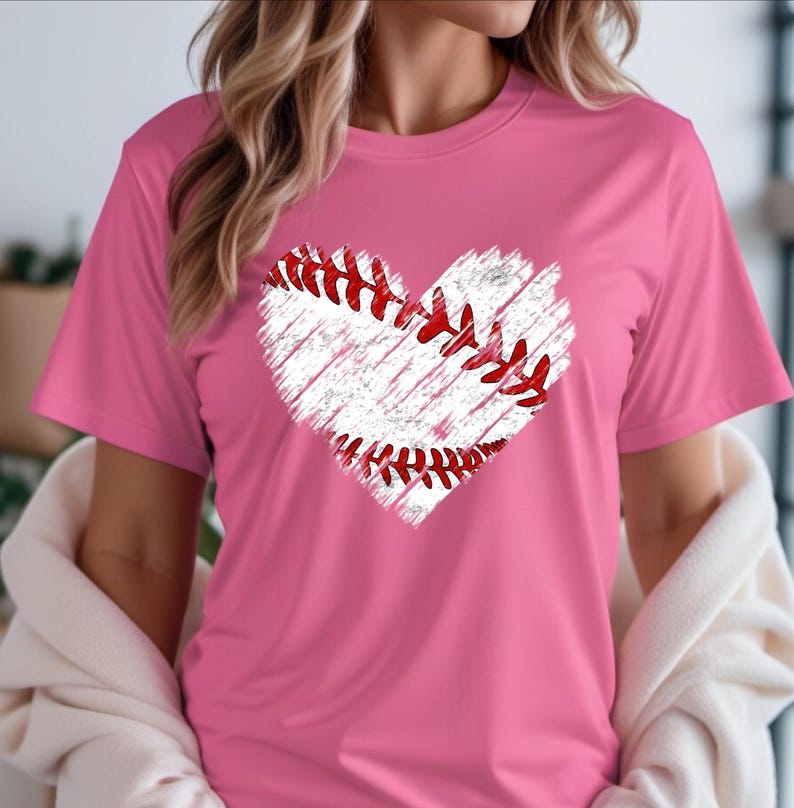 Puede incluir: Una camiseta rosa con un gr&aacute;fico de b&eacute;isbol blanco en forma de coraz&oacute;n. El b&eacute;isbol tiene costuras rojas.