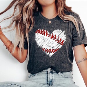Puede incluir: Una camiseta gris con un gr&aacute;fico de b&eacute;isbol en forma de coraz&oacute;n blanco. El b&eacute;isbol tiene costuras rojas.