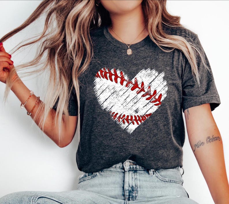 Puede incluir: Una camiseta gris con un gr&aacute;fico de b&eacute;isbol en forma de coraz&oacute;n blanco. El b&eacute;isbol tiene costuras rojas.