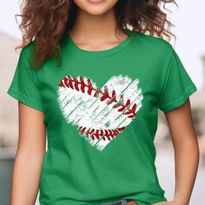 Puede incluir: Una camiseta verde con un gr&aacute;fico de b&eacute;isbol blanco en forma de coraz&oacute;n. El b&eacute;isbol tiene costuras rojas.