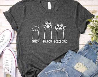 Può includere: Una maglietta grigia scura con una grafica bianca di un'impronta di zampa, un'impronta di zampa con artigli e un'impronta di zampa con un cerchio in cima. Il testo "Rock Paper Scissors" è stampato sotto la grafica.