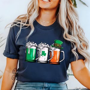以下が含まれることがあります： 紺色のTシャツに、ビールジョッキ3つのグラフィックがプリントされています。ジョッキは緑、白、オレンジのビールで満たされています。オレンジのビールのジョッキには、緑の妖精の帽子が乗っています。