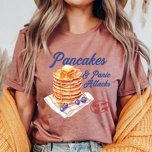 Może przedstawiać: Koszulka w kolorze malwy z grafiką stosu naleśników z syropem i borówkami. Na koszulce nadrukowany jest tekst "Pancakes & Panic Attacks" w kolorze niebieskim. Tekst "Buy one get one" jest nadrukowany na białym kółku w kolorze czerwonym.