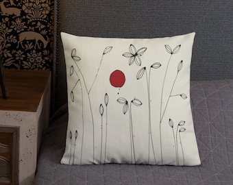 May include: A white throw pillow with a black and red minimalist design featuring stylized flowers and a red circle. The pillow is on a gray couch.