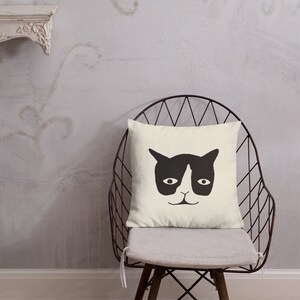 May include: A square, cream-colored throw pillow featuring a minimalist black cat face design. The pillow is placed on a metal chair with a gray cushion, set against a textured gray wall.