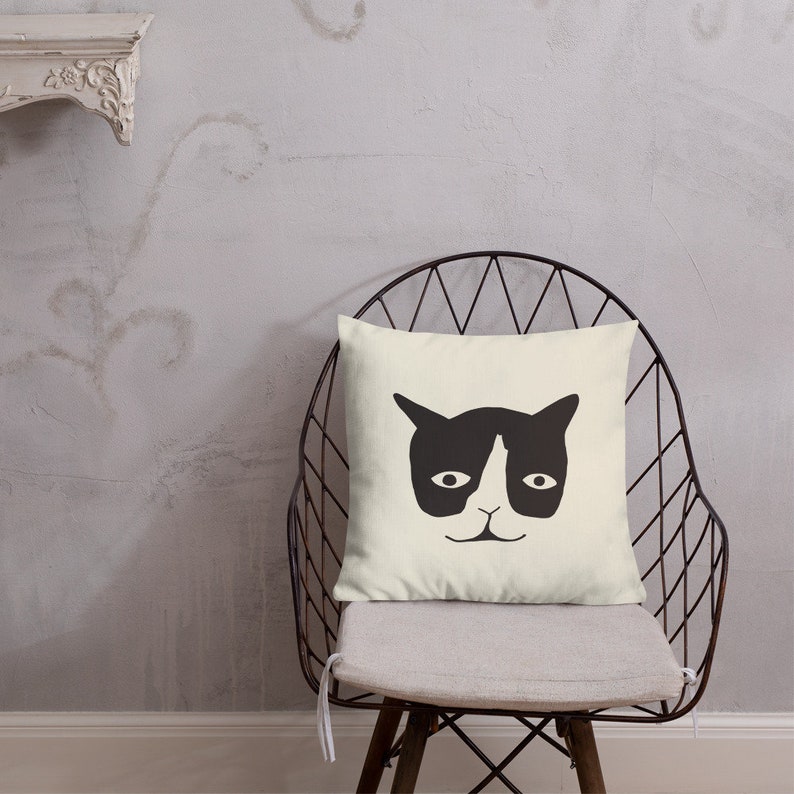 May include: A square, cream-colored throw pillow featuring a minimalist black cat face design. The pillow is placed on a metal chair with a gray cushion, set against a textured gray wall.