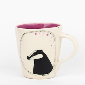 May include: A ceramic mug with a cream-colored exterior featuring a hand-drawn black and white badger illustration inside a thin oval frame. The interior of the mug is a vibrant magenta, and the exterior is accented with small purple dots.