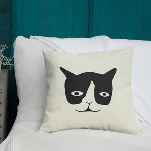 May include: Square throw pillow with a black and white cat face design. The cat's face is centered on the pillow, with large eyes, pointed ears, and a small nose and mouth. The pillow is on a white bed.