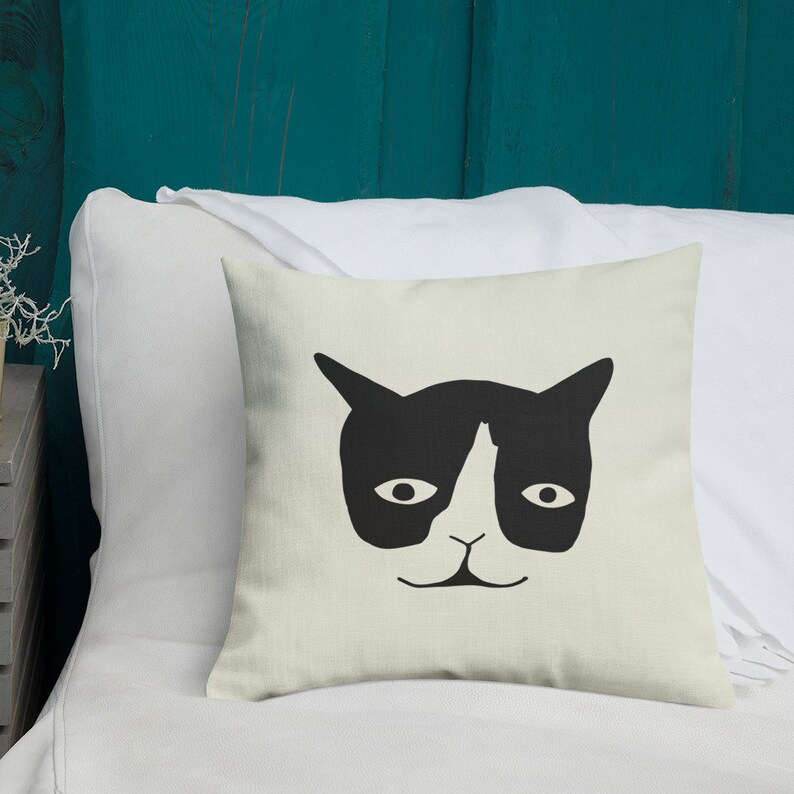 May include: Square throw pillow with a black and white cat face design. The cat's face is centered on the pillow, with large eyes, pointed ears, and a small nose and mouth. The pillow is on a white bed.