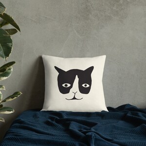 May include: Cream-colored square throw pillow featuring a black cat face design. The cat's face has a black mask, ears, and a nose, with two oval eyes and a small mouth. The pillow is on a blue blanket.