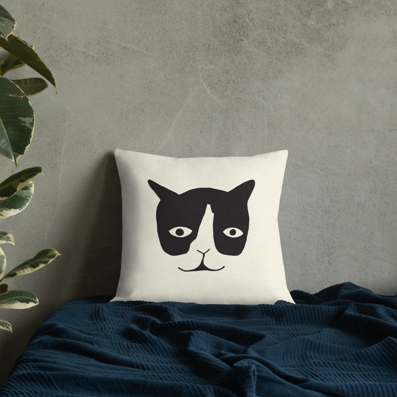 May include: Cream-colored square throw pillow featuring a black cat face design. The cat's face has a black mask, ears, and a nose, with two oval eyes and a small mouth. The pillow is on a blue blanket.