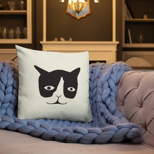 May include: Square throw pillow with a black and white cat face design. The cat's face is centered on the pillow, with a black head and ears, white face, and oval eyes. The pillow is on a pink couch with a blue blanket.