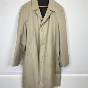 Op de afbeelding: Een beige trenchcoat hangt aan een houten kleerhanger. De jas heeft een klassiek ontwerp met een reverskraag, een knoopsluiting en een knielange zoom. De binnenvoering is donker. De jas is gemaakt van een gladde, duurzame stof.