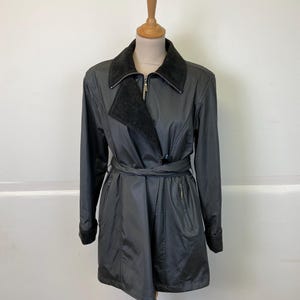 Op de afbeelding: Een zwarte trenchcoat met een donkergrijze kraag en bijpassende riem. De jas heeft een ritssluiting en twee zijzakken. De jas wordt gepresenteerd op een houten mannequin.