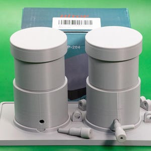 May include: A gray, 3D-printed device with two cylindrical components, each topped with a white lid. The device sits on a rectangular base with additional small parts. A box with the text "Mitron" is in the background.