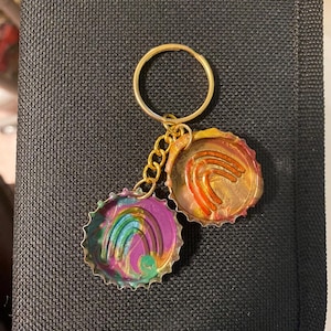 May include: A gold-coloured keyring with two bottle cap charms. One charm features a rainbow design in purple, blue, green, and pink. The other charm has orange and red swirls. The keyring is attached to a black surface.