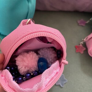 May include: A small pink zippered pouch with a blue pom-pom, a blue bead, and a purple beaded necklace inside. The pouch is open and the contents are visible.