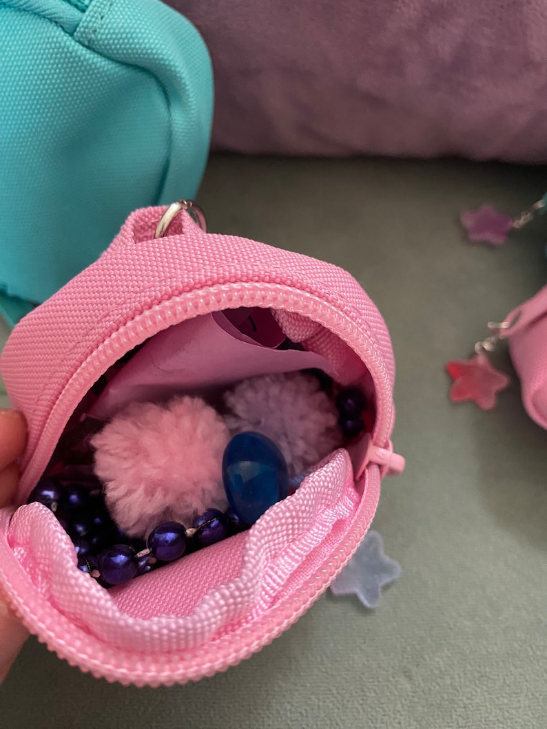 May include: A small pink zippered pouch with a blue pom-pom, a blue bead, and a purple beaded necklace inside. The pouch is open and the contents are visible.