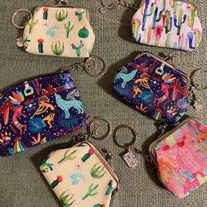 May include: Six different small coin purses with keychains. The purses are made of fabric and have various colorful patterns. The patterns include cacti, flowers, and geometric designs. The keychains are silver and have a small charm attached.
