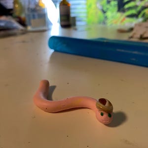 May include: A pink worm-shaped figurine with a smiling face and a small hat. The worm has green eyes and a light brown hat. The figurine is on a white surface.