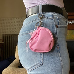May include: A pink zippered pouch with a silver chain attached to a belt loop. The pouch is hanging from the belt loop of a pair of blue jeans.