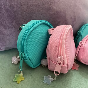 May include: Three small, round backpacks with zipper closures. The backpacks are made of a soft, canvas-like material. The backpacks are in teal, pink, and white. Each backpack has a star-shaped charm attached to the zipper pull.