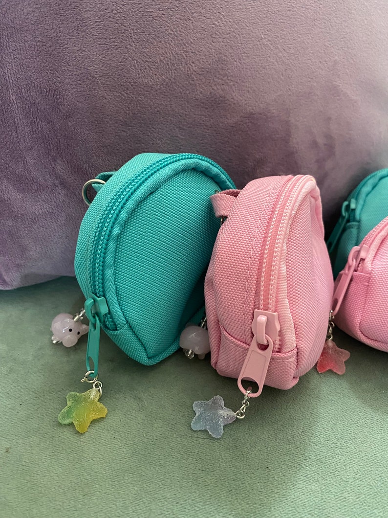 May include: Three small, round backpacks with zipper closures. The backpacks are made of a soft, canvas-like material. The backpacks are in teal, pink, and white. Each backpack has a star-shaped charm attached to the zipper pull.