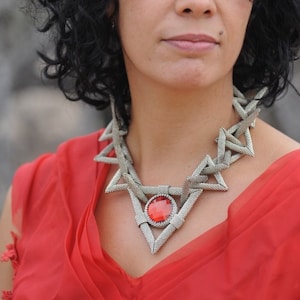 以下が含まれることがあります： 黒髪をカールさせた女性が赤いドレスを着て、大きな赤い宝石のペンダントが付いた銀のネックレスをしています。