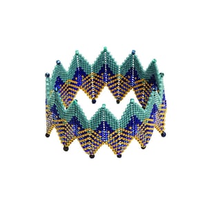 May include: A beaded bracelet featuring a geometric pattern in shades of blue, green, and gold. The bracelet is crafted from small, closely woven beads and boasts a distinctive, contemporary design.