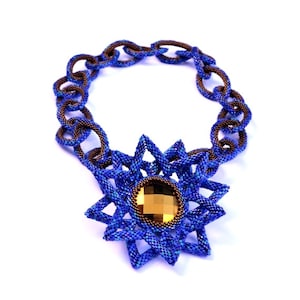 以下が含まれることがあります： 大きな金茶色の宝石を中央に配した、星型のペンダントが付いたビーズネックレス。 ネックレスは青と茶色のビーズのチェーンで構成されています。 ジュエリーは白い背景に配置されています。